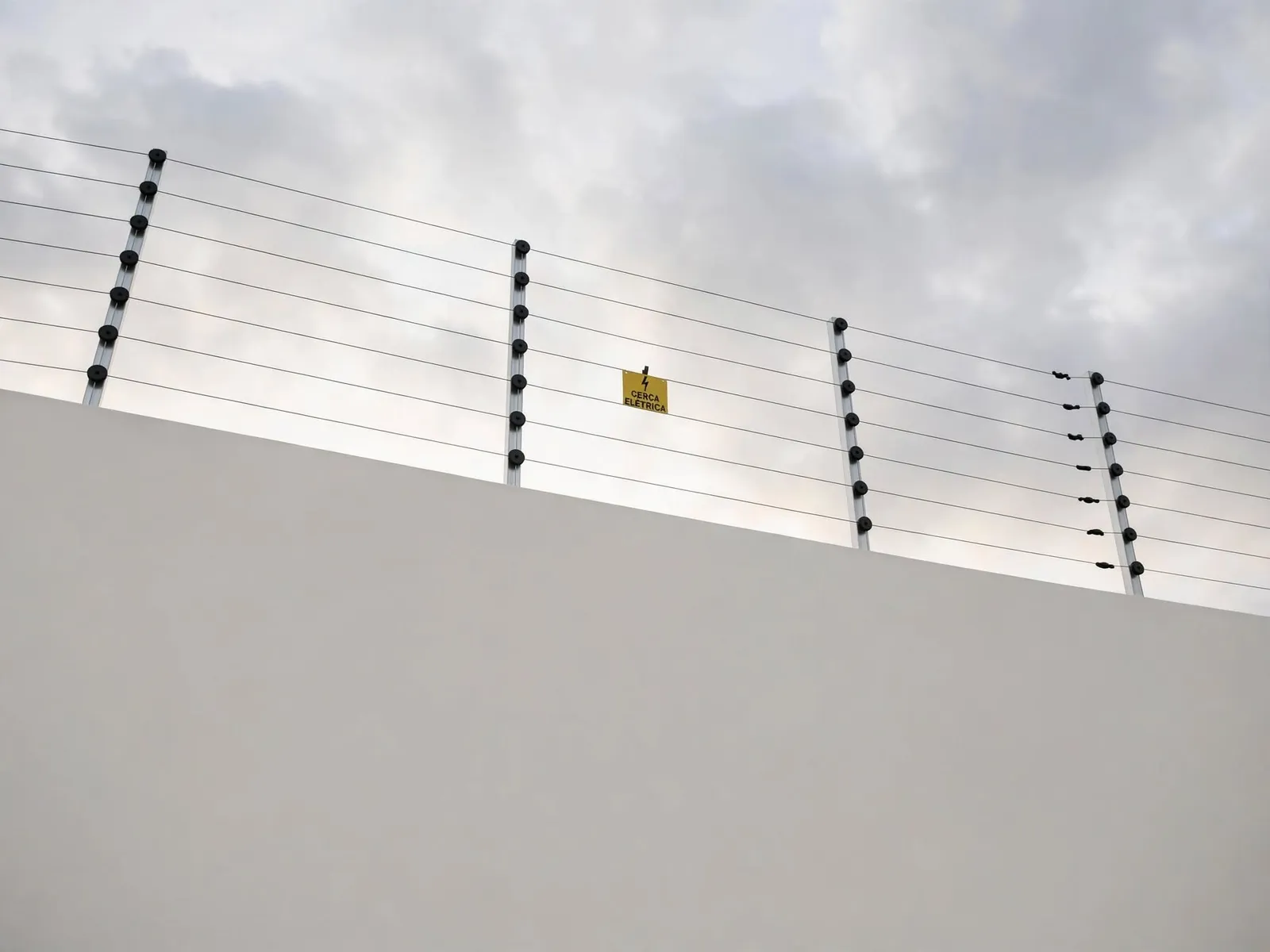 Cerca elétrica no alto de um muro, com placa amarela de aviso